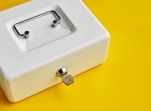Close up of small white closed cashbox or money box with key in lock on yellow background with copy space.