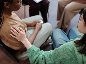 Young brunette woman in casual attire keeping hand on shoulder of upset African American girl while comforting her during session