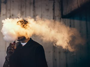 A dark and high contrast image of a young adult male has a cloud of nicotine vapor "smoke" pouring from his mouth.  Vape smoking, or "vaping" is growing in popularity, as well as falling under stricter state and governmental regulations.  Horizontal image with copy space.